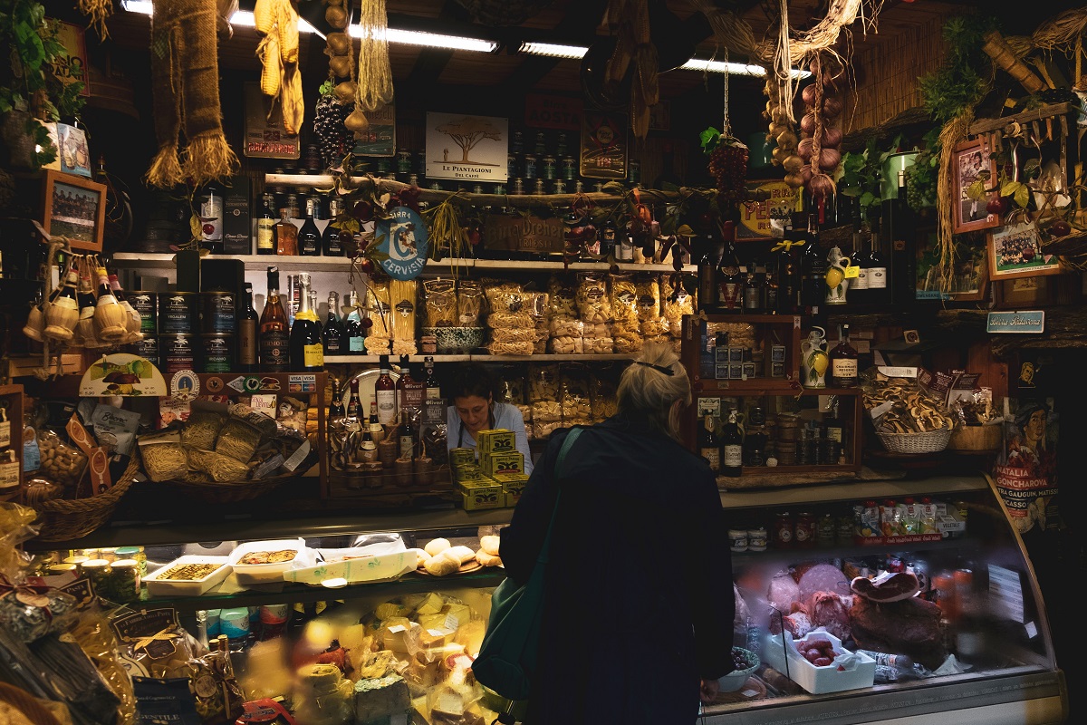 Una mujer de mediana edad compra en un puesto de comida lleno de carne, queso, pan y productos envasados ​​dentro de un mercado en Florencia.