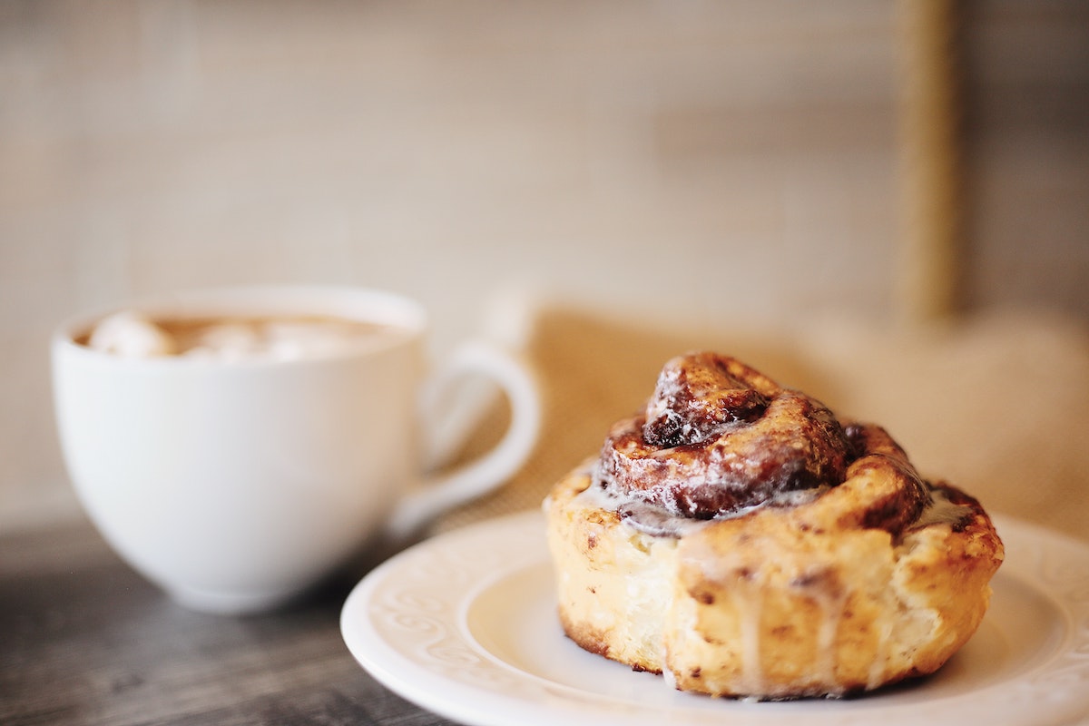 Un rollo de canela en un plato blanco junto a una taza de café blanca.
