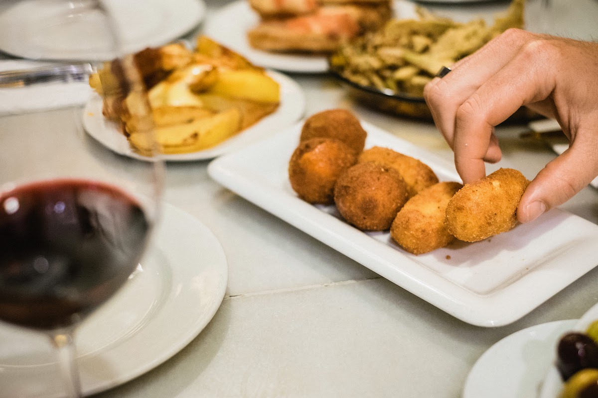 La mano de una persona tomando una croqueta frita de un pequeño plato blanco, con una copa de vino tinto visible en primer plano.