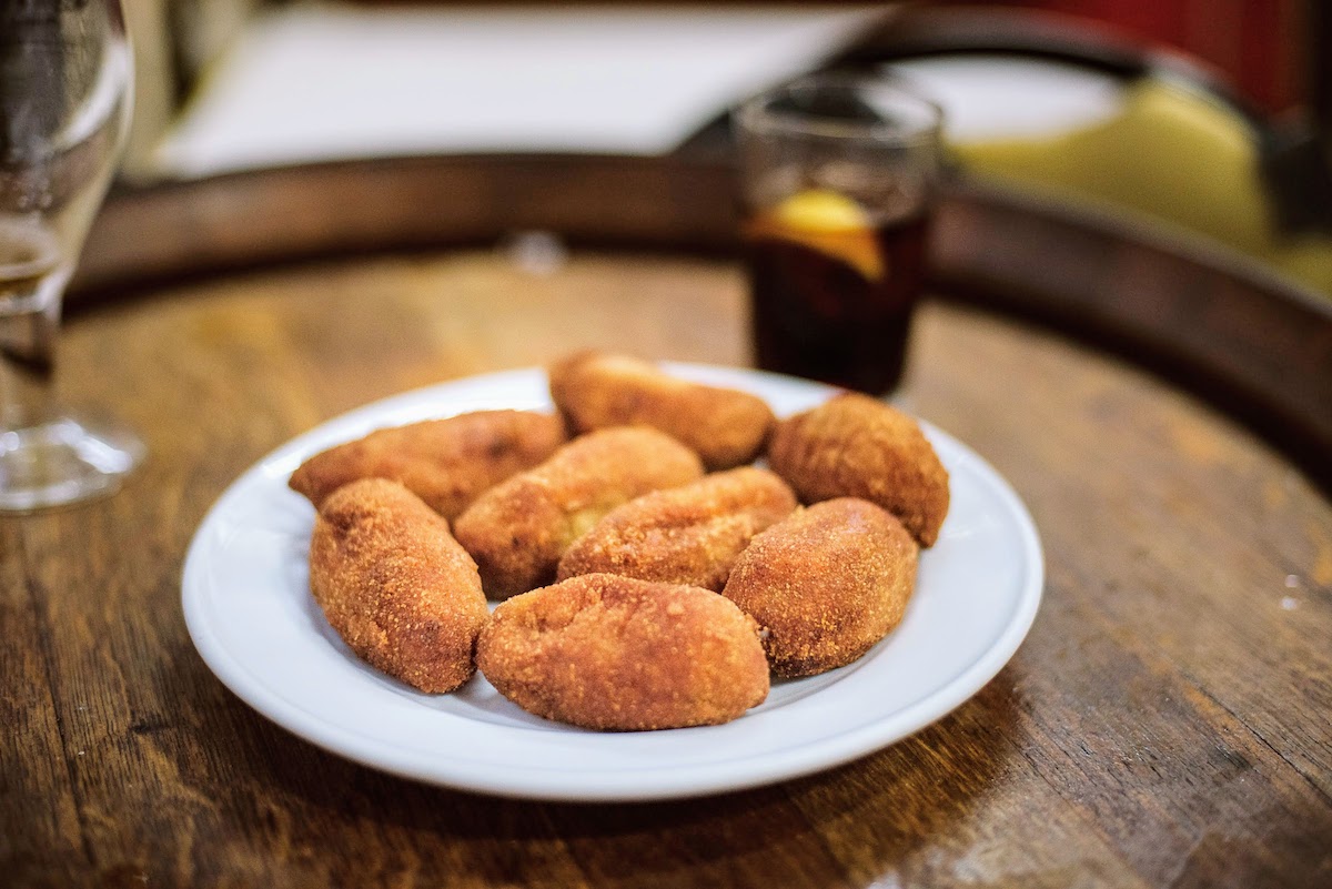 Unas croquetas en un plato blanco, con una copa de vermut visible al fondo.