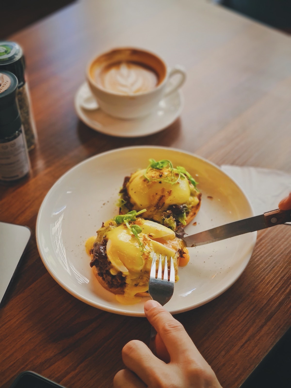 Una persona cortando huevos benedictinos con una taza de café en la mesa al fondo