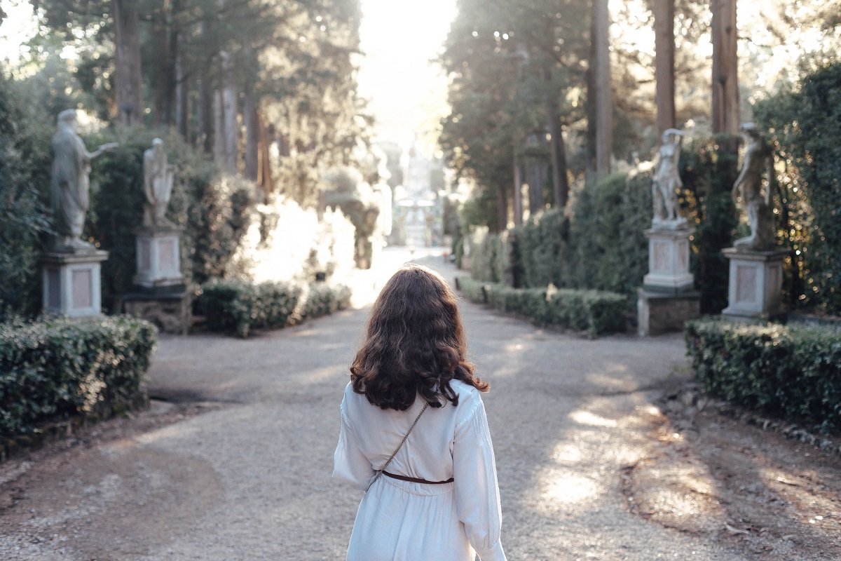 Una mujer con un vestido blanco camina por un bulevar arbolado y flanqueado por estatuas.