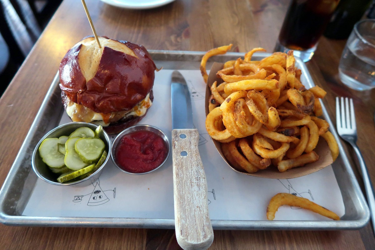Una pequeña hamburguesa en una bandeja con una gran porción de papas fritas rizadas, pepinillos y ketchup con un gran cuchillo de madera