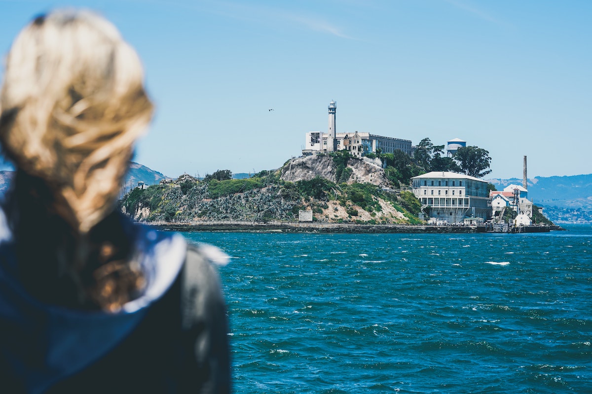 Una persona con el pelo azotado por el viento en primer plano mira a través de la bahía hacia la isla de Alcatraz, un lugar aterrador para visitar en San Francisco.