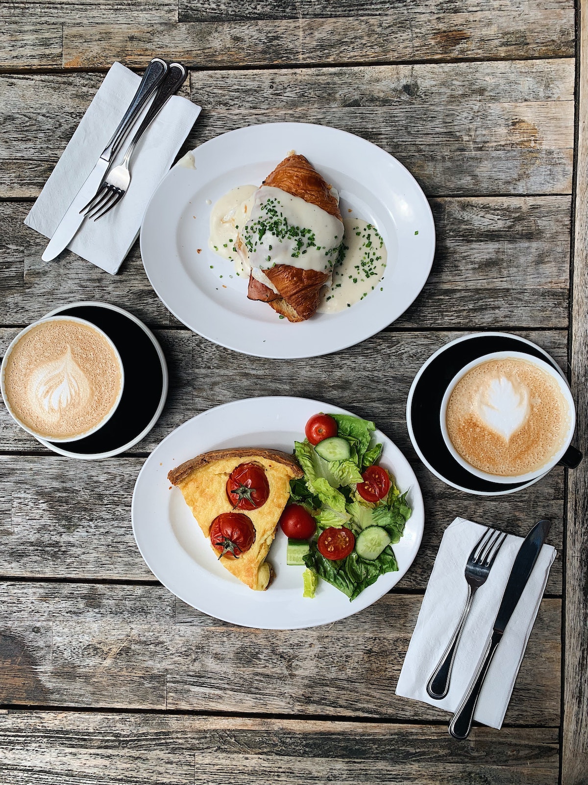 Plano de una mesa de madera con dos capuchinos y un plato con quiche y ensalada, el otro con un croissant