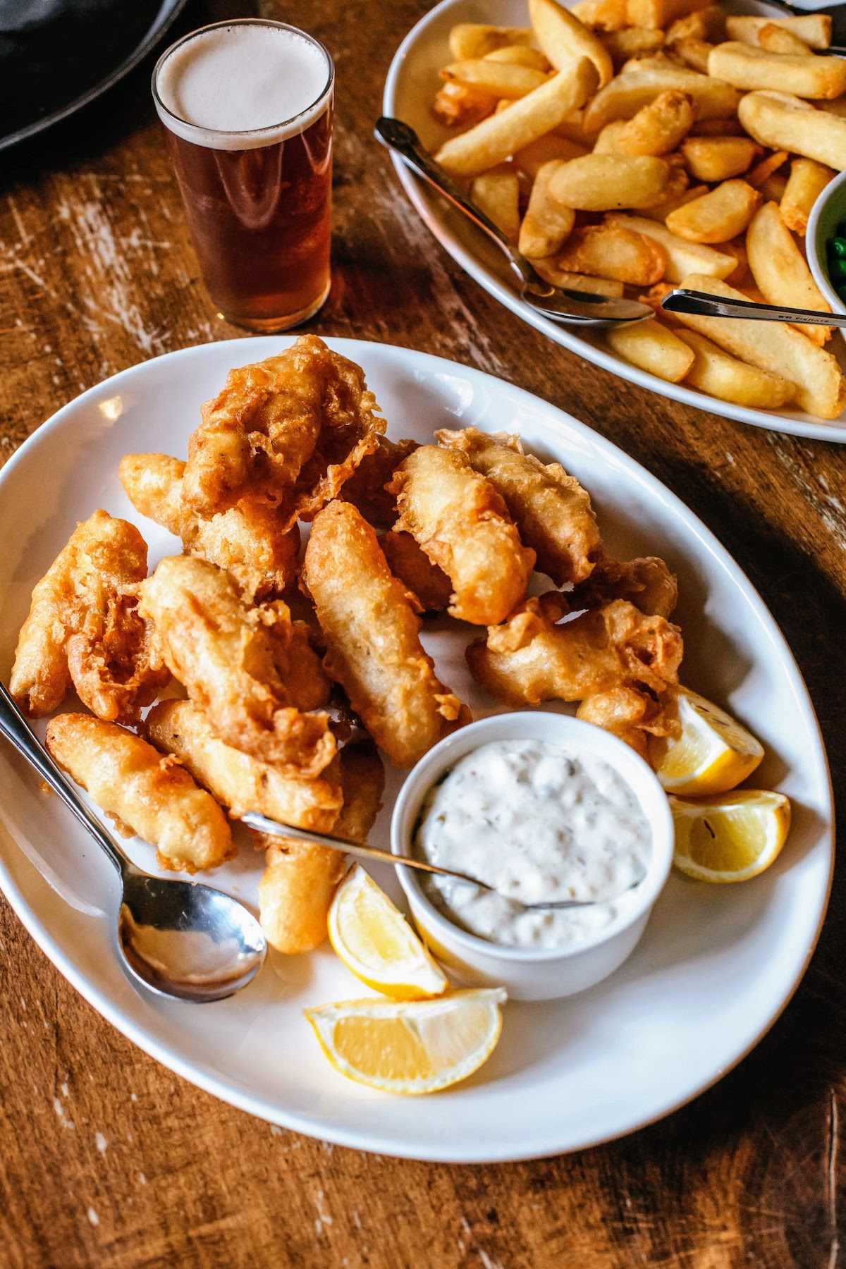 Trozos de pescado frito servido en un plato blanco con salsa tártara y gajos de limón con un plato de patatas fritas de fondo.