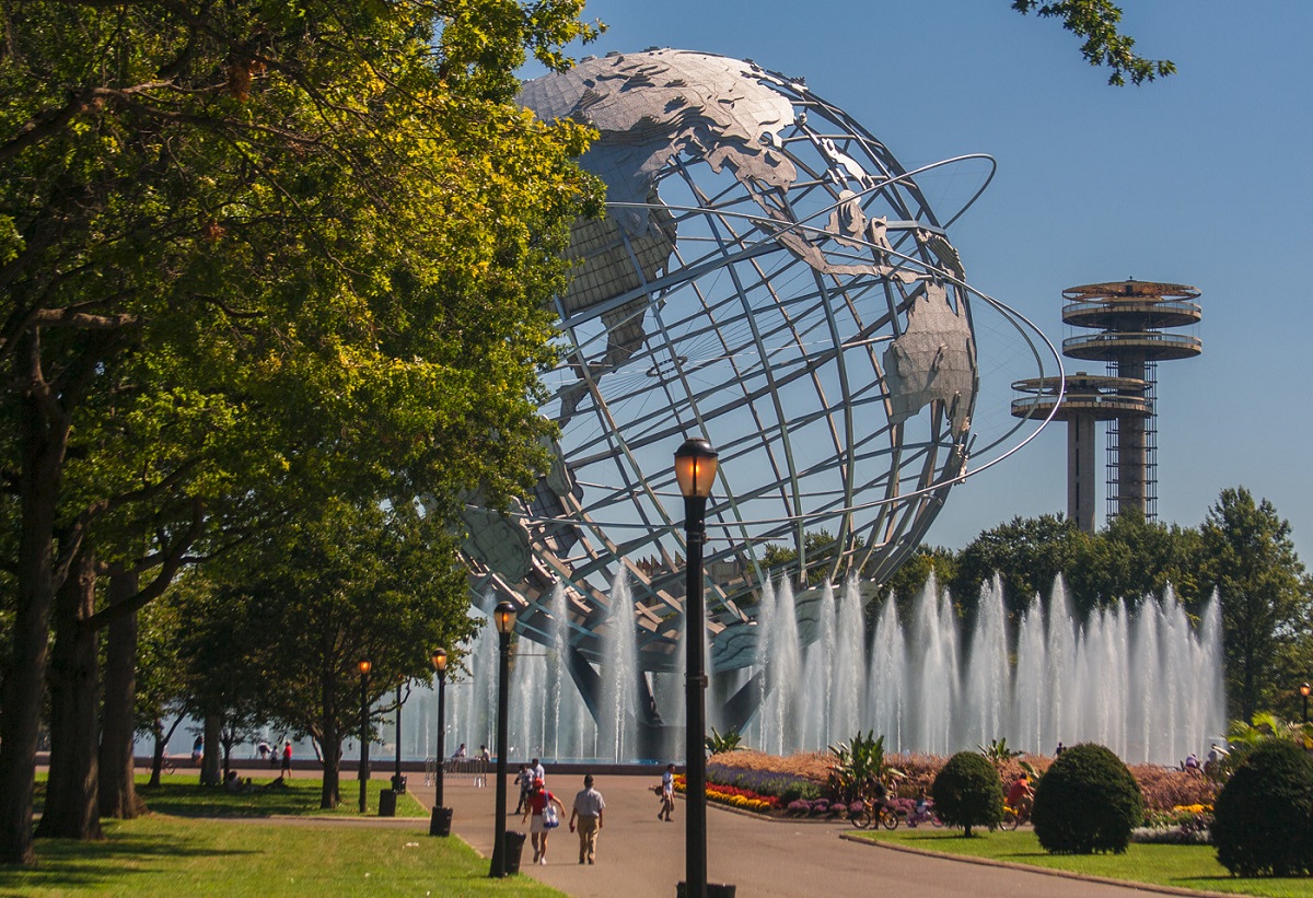 Una esfera plateada gigante con forma de tierra emerge de detrás de árboles y fuentes de agua en un parque de Nueva York.