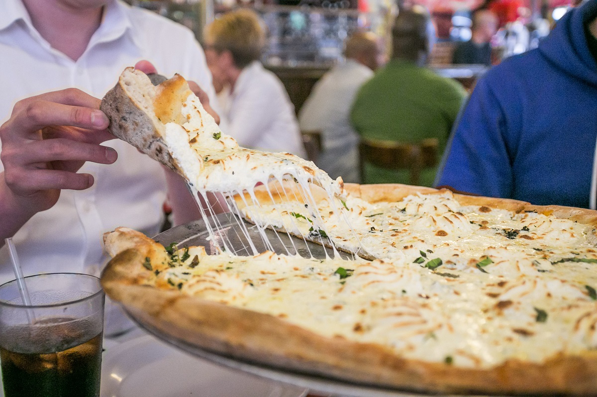 Un hombre sacando una porción de pizza blanca de una sartén con tiras de queso que la conectan a la pizza principal
