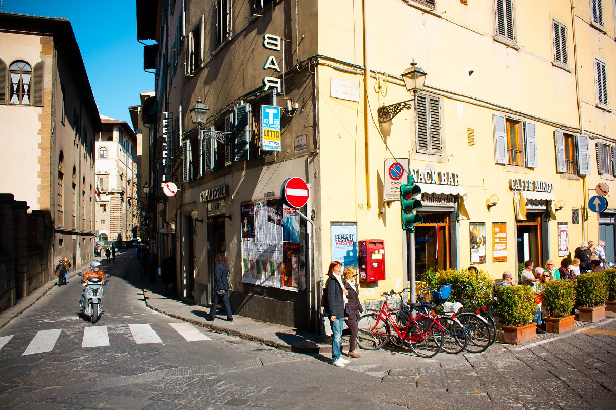 Dos mujeres esperan afuera de un edificio al sol con sus bicicletas en una calle de Florencia.