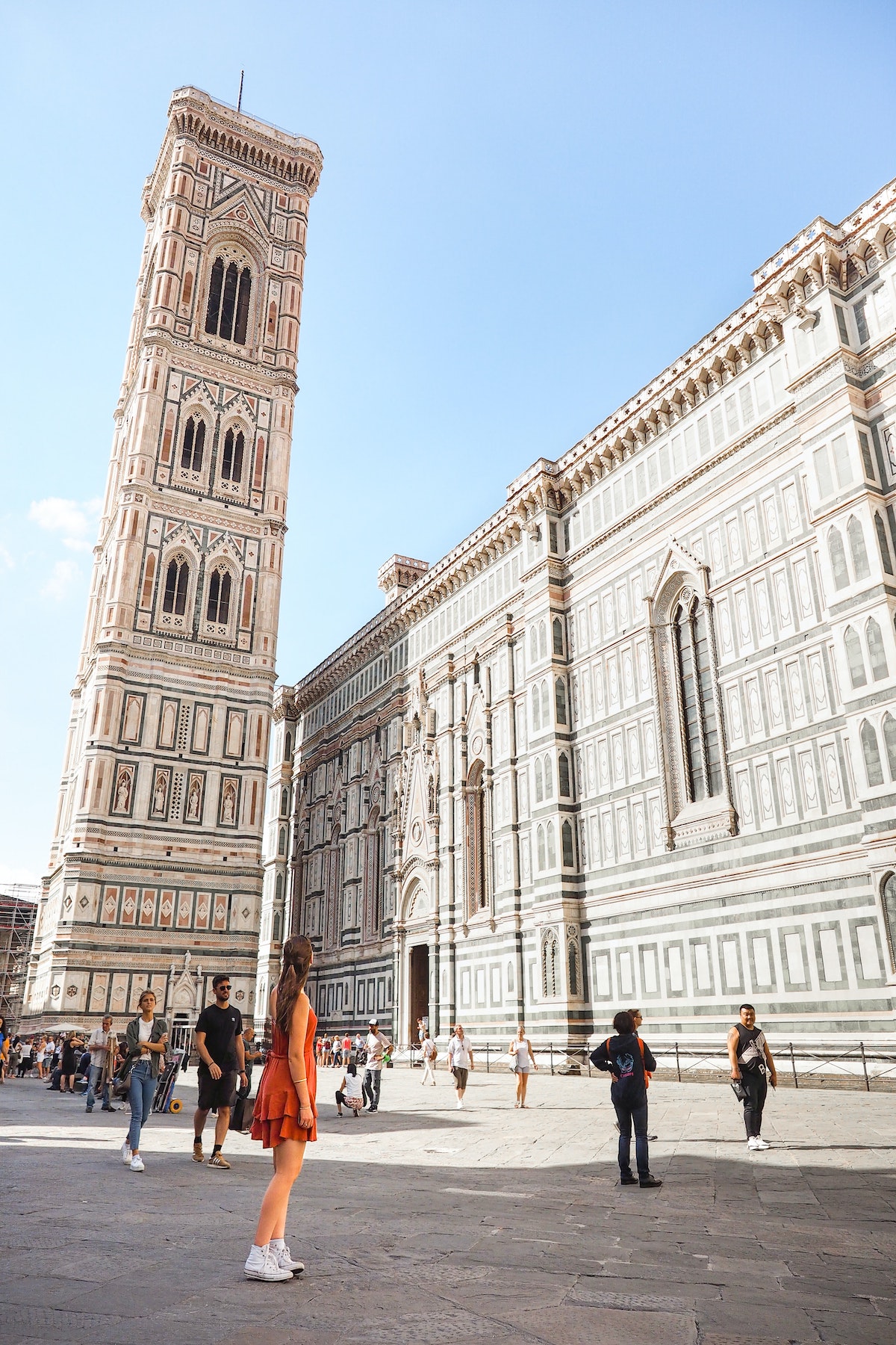 Una mujer con un vestido rojo parada frente al campanario de la catedral en una gran plaza abierta