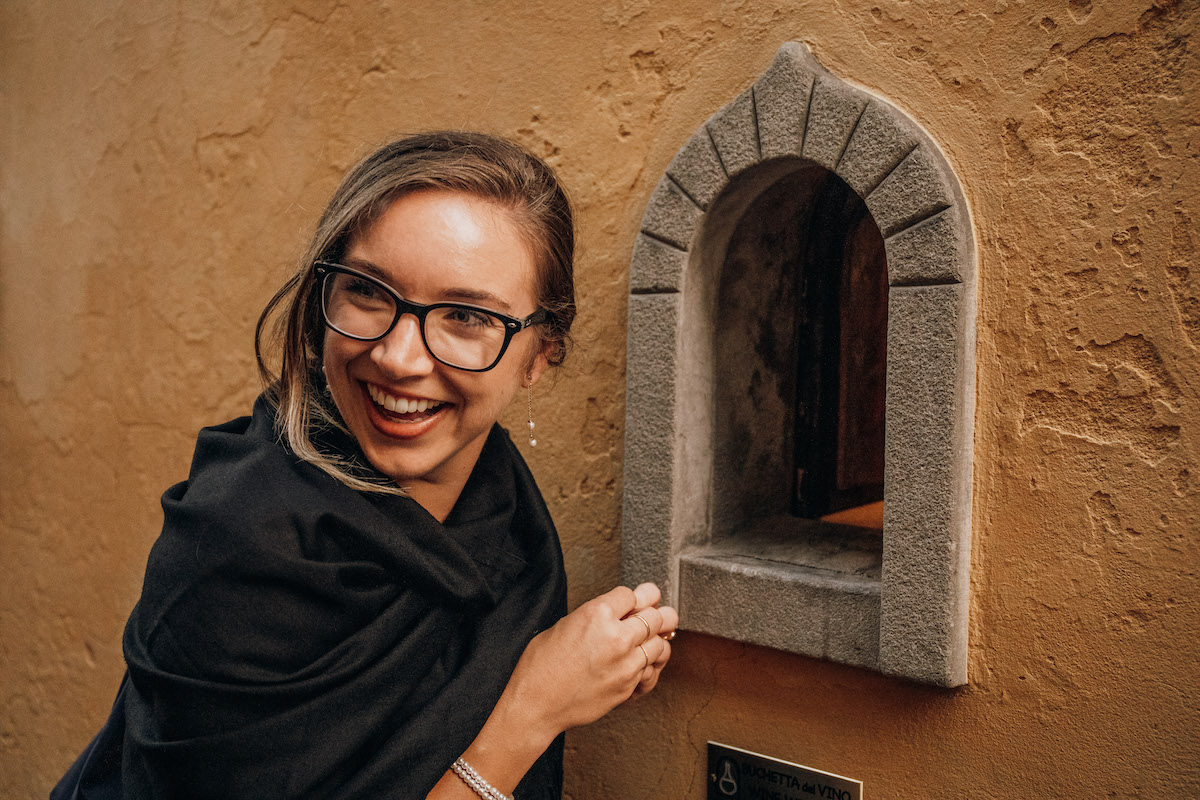 Una mujer con gafas sonriendo frente a una pequeña abertura en forma de arco en un muro de piedra