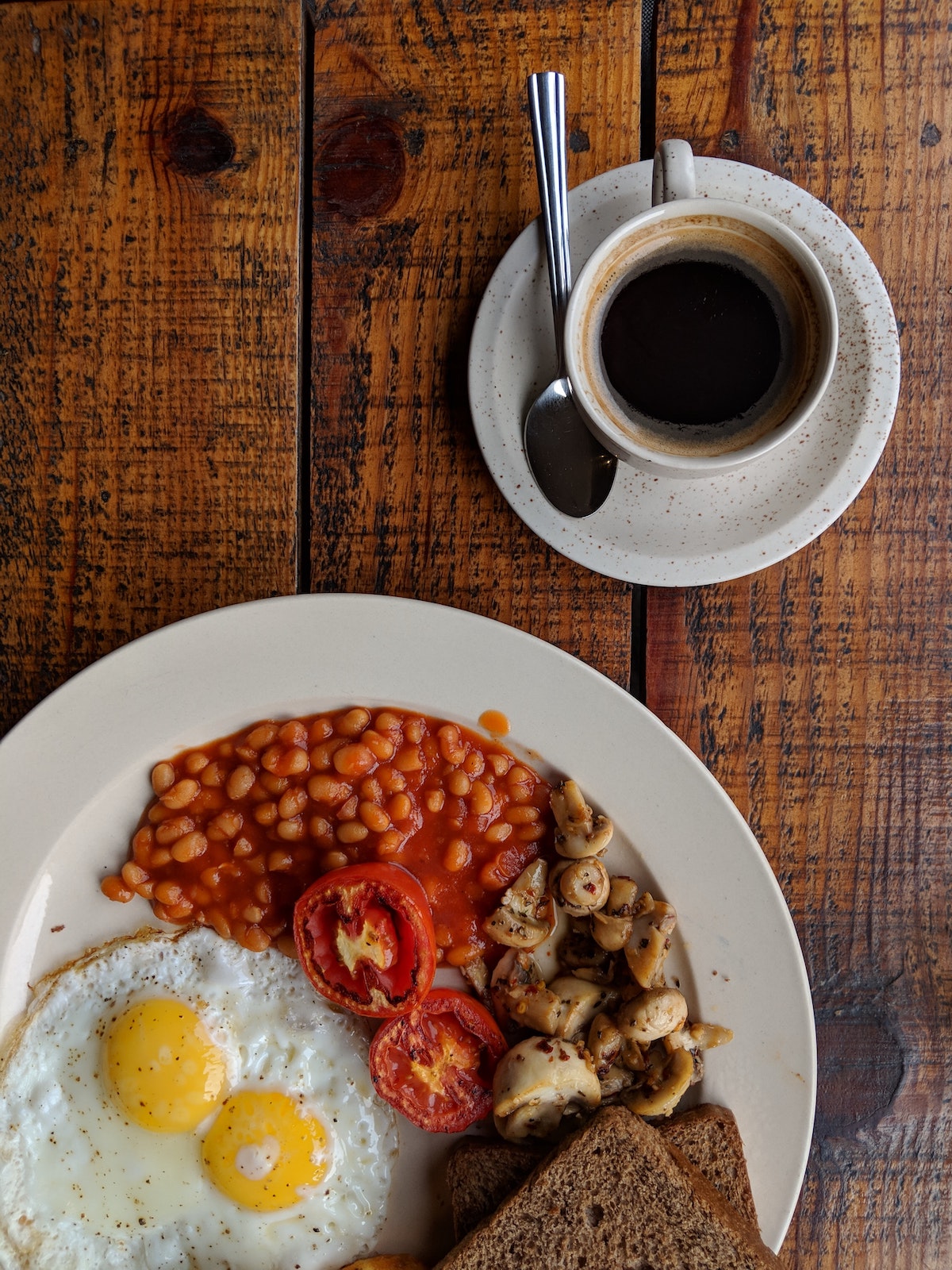 Fotografía superior de un desayuno inglés completo que incluye frijoles, huevos, champiñones y tomates en un plato blanco junto a una taza de café