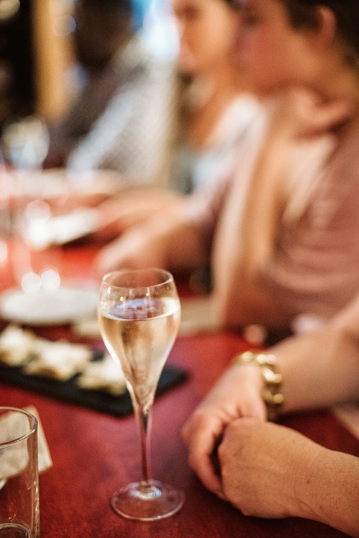 Una copa de vino espumoso en una mesa roja rodeada de gente y otros platos