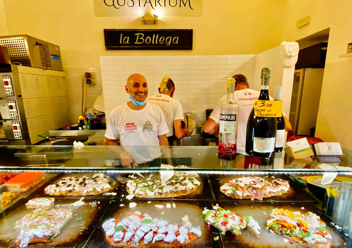Un hombre sonriendo detrás de un mostrador con algunas pizzas en exhibición