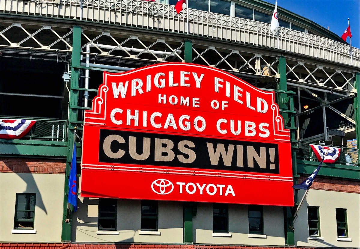 Un gran letrero rojo dice "Wrigly Field Home of Chicago Cubs" en letras blancas. Las luces LED debajo dicen "CUBS WIN!" Ir a un juego de los Cachorros en Chicago con niños es genial.