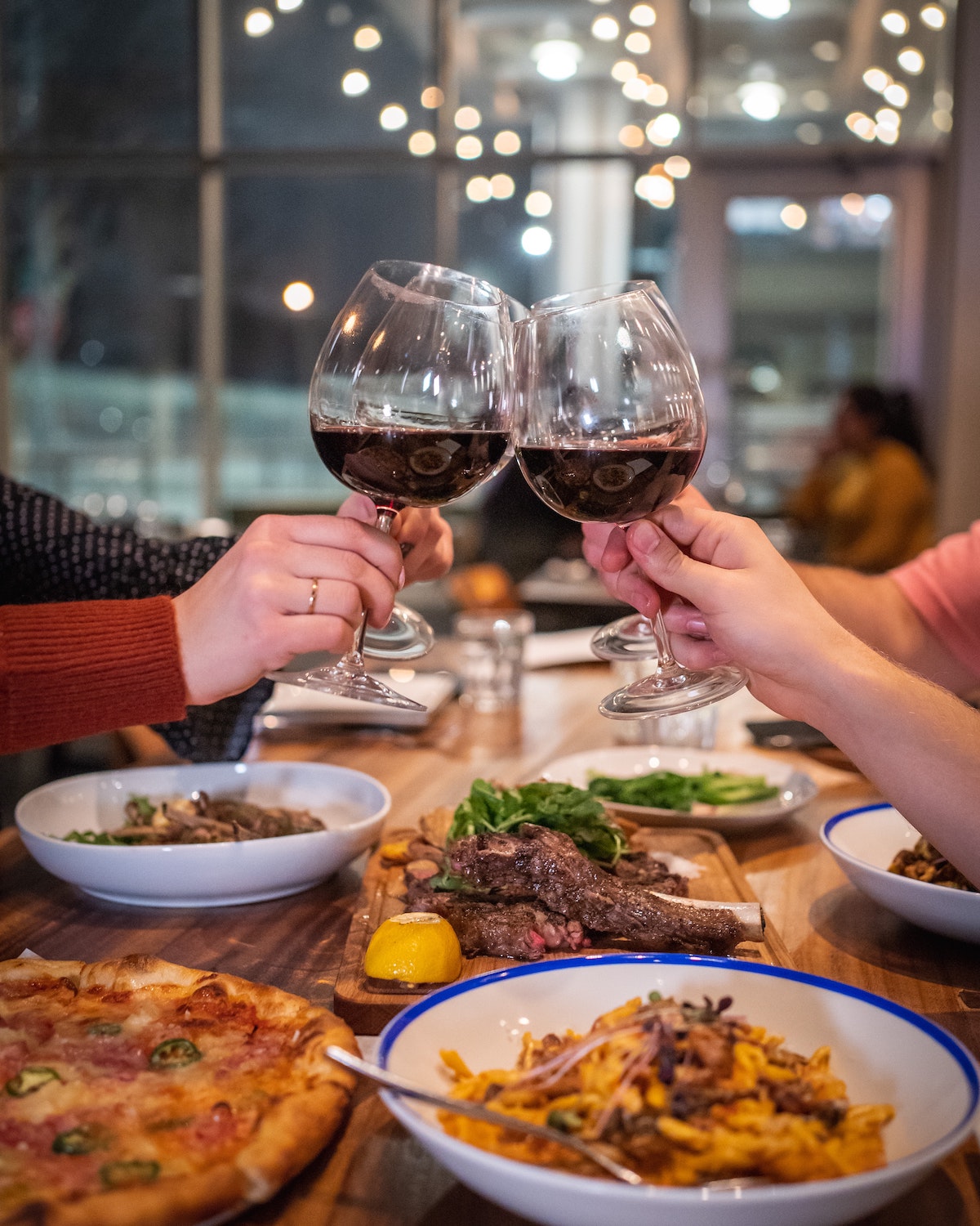 Personas brindando con copas de vino tinto sobre una mesa llena de pizza, pasta y bistec