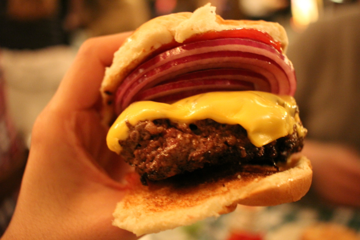 La mano de una persona sosteniendo una hamburguesa con queso con una cebolla roja.