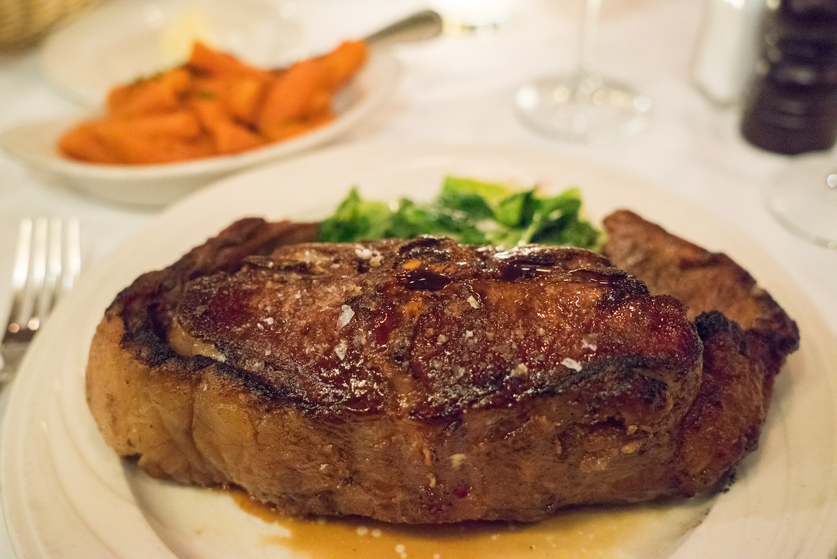 Un trozo de bistec espolvoreado con sal gruesa en un plato blanco con una porción de verduras al fondo