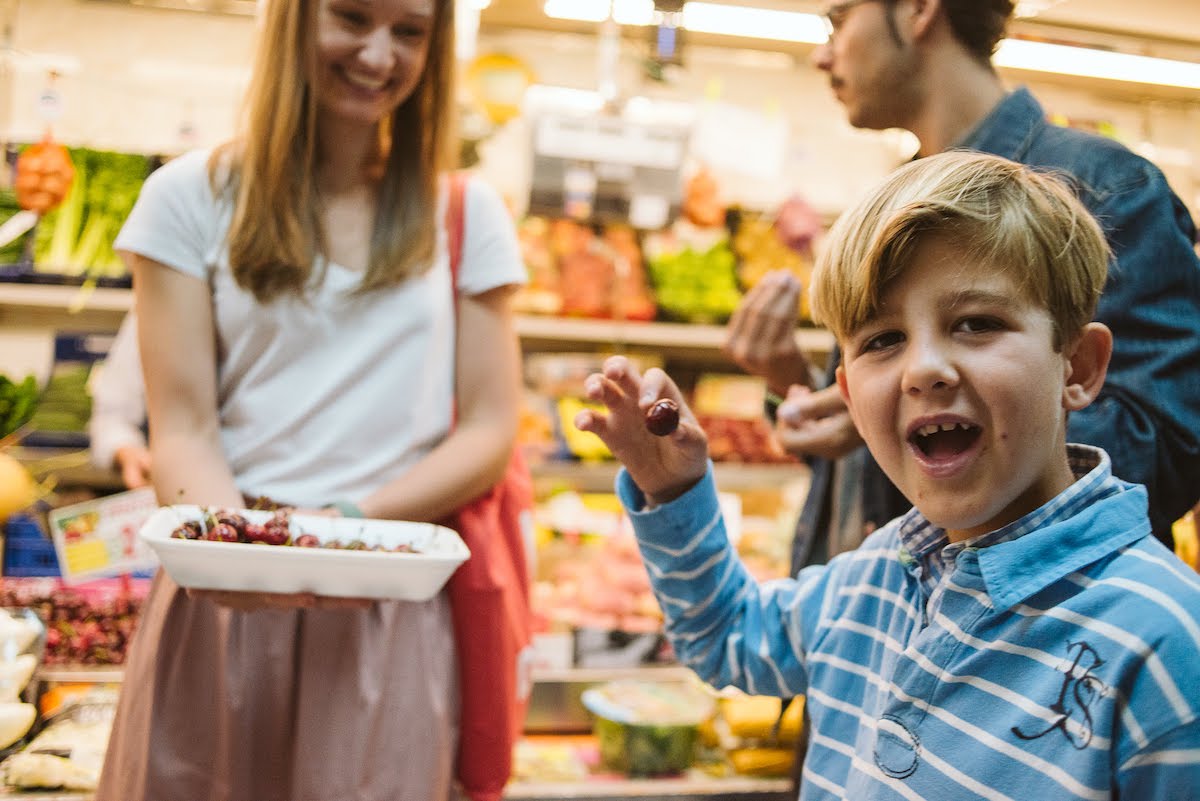Un niño sonriendo mientras come cerezas de una bandeja blanca frente a un puesto en el mercado