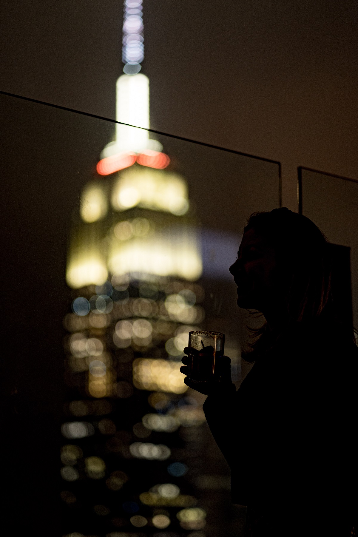 Una mujer bebe un cóctel por la noche mientras las luces de Manhattan iluminan el fondo oscuro de la foto.
