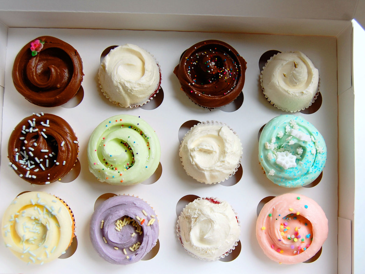 Fotografía cenital de una docena de cupcakes de diferentes colores en una caja de panadería blanca