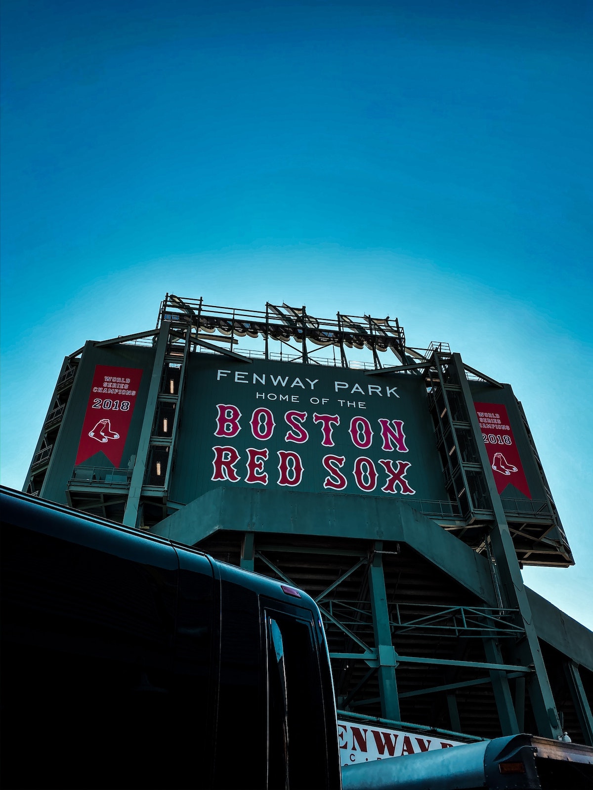Un letrero verde gigante con letras rojas y blancas dice "Fenway Park, hogar de los Medias Rojas de Boston" contra un cielo azul vibrante