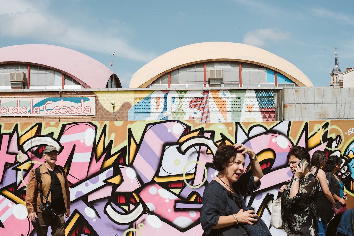 Gente caminando frente a coloridos murales de arte callejero en las paredes exteriores de un gran edificio de mercado.