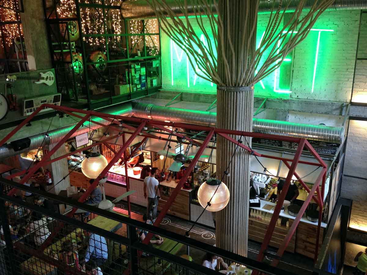 Una vista desde el segundo piso de un mercado gastronómico con luces de neón y una decoración moderna.