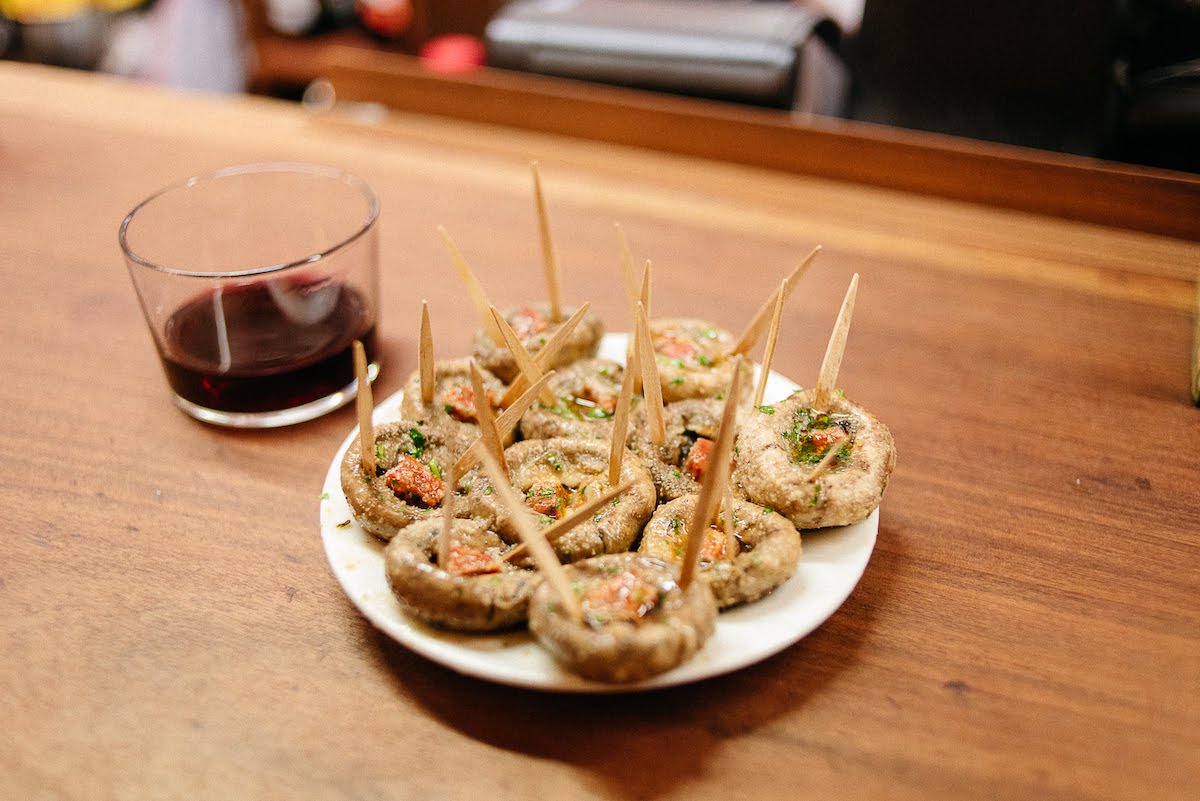 Un plato de champiñones a la plancha con chorizo ​​junto a una copa corta de vino tinto.