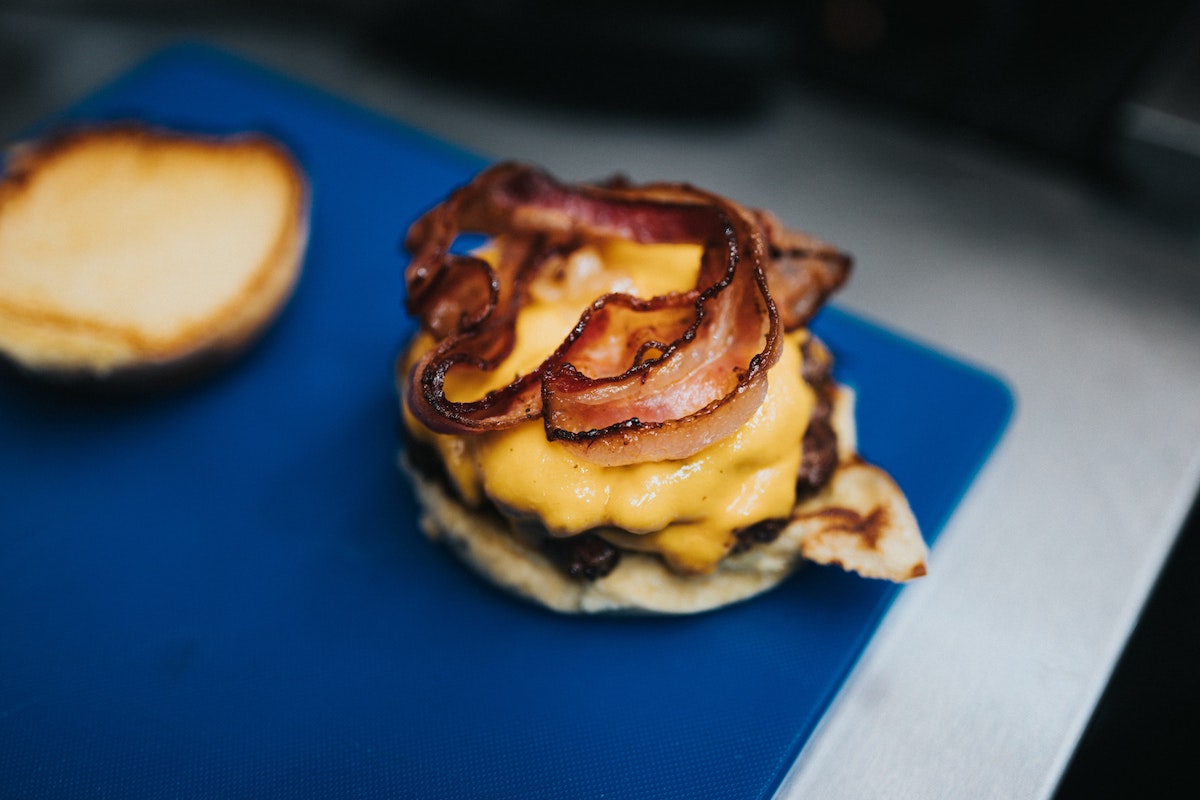 Una hamburguesa con queso y tocino se sienta en una tabla de cortar azul con la parte superior hacia un lado para que podamos ver el tocino crujiente y el queso amarillo derretido 