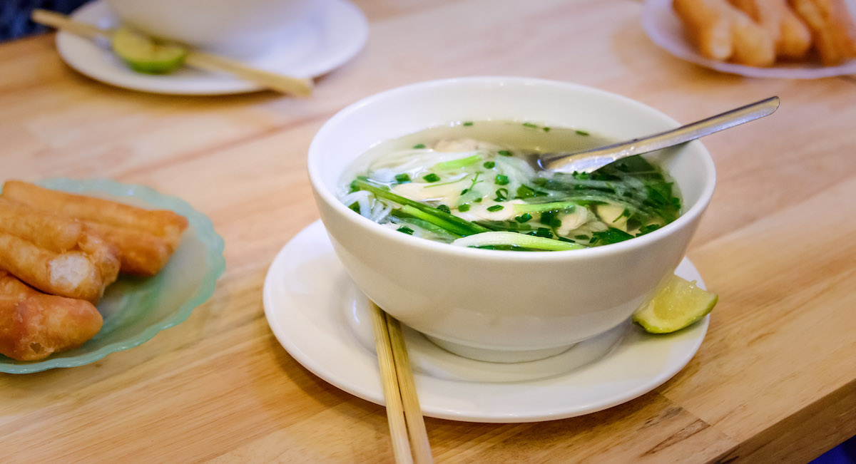 Un plato de sopa de fideos con cebollino y vegetales verdes en un plato blanco junto a una rodaja de cítricos y un par de palillos