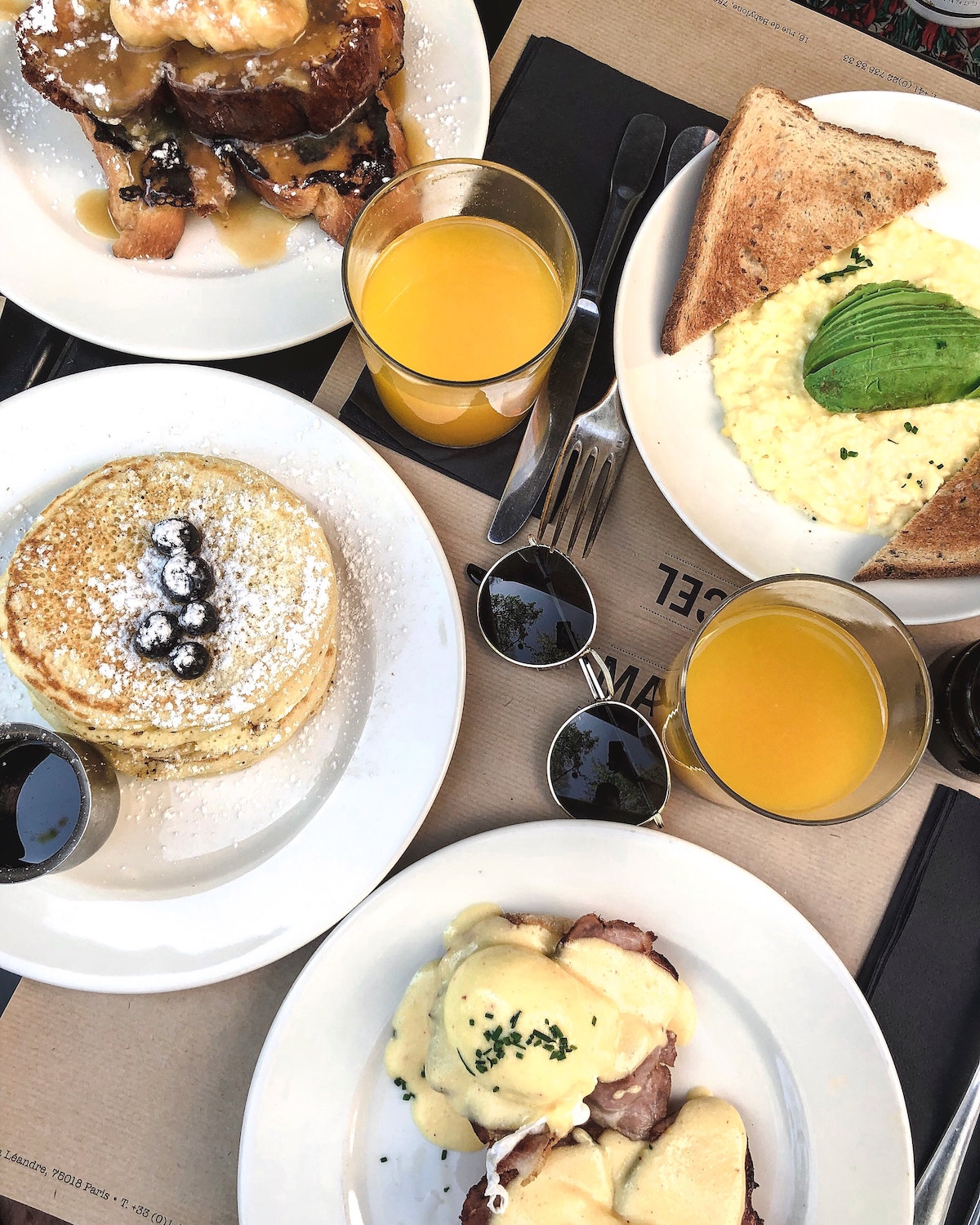 Fotografía cenital de artículos para el desayuno en una mesa que incluye panqueques, huevos benedictinos, tostadas y dos vasos de jugo de naranja, con un par de lentes de sol en el medio