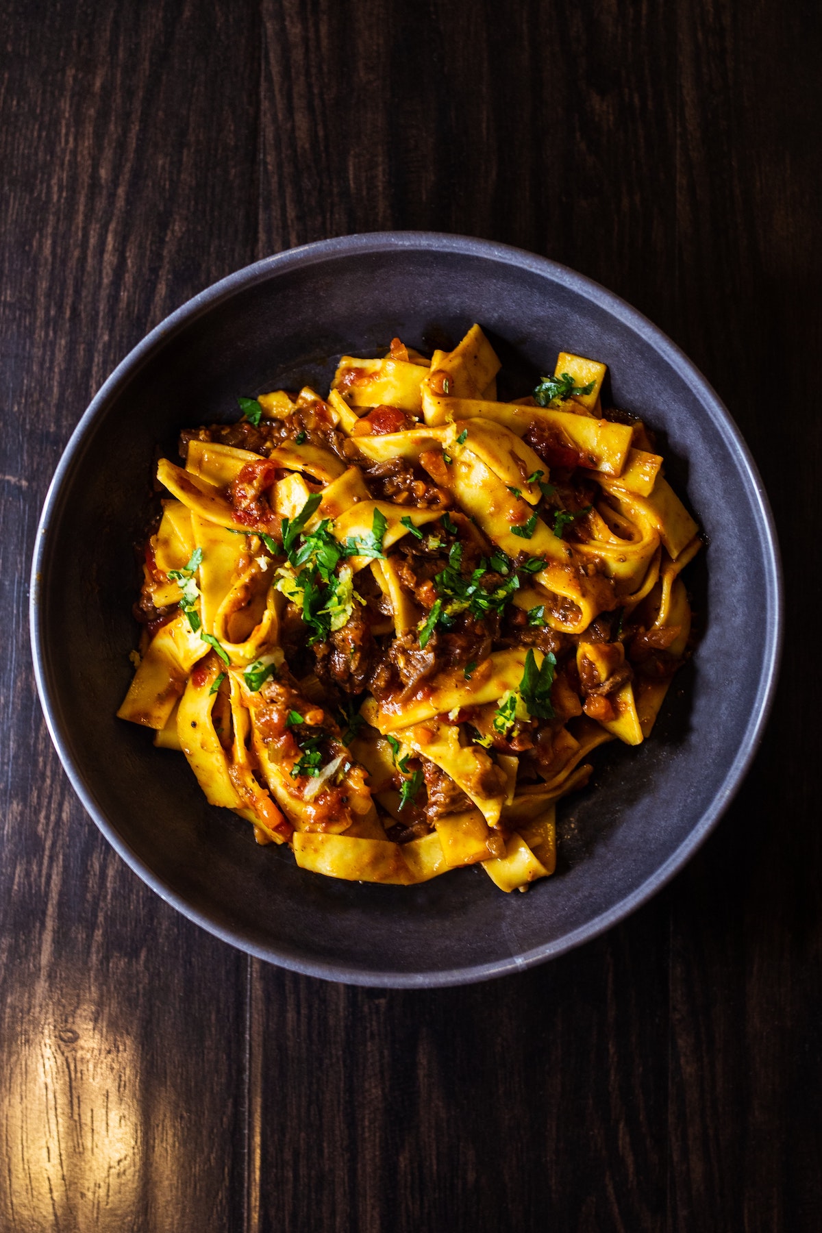 Fotografía cenital de pasta larga y ancha con salsa de carne y hierbas en un bol sobre un fondo oscuro
