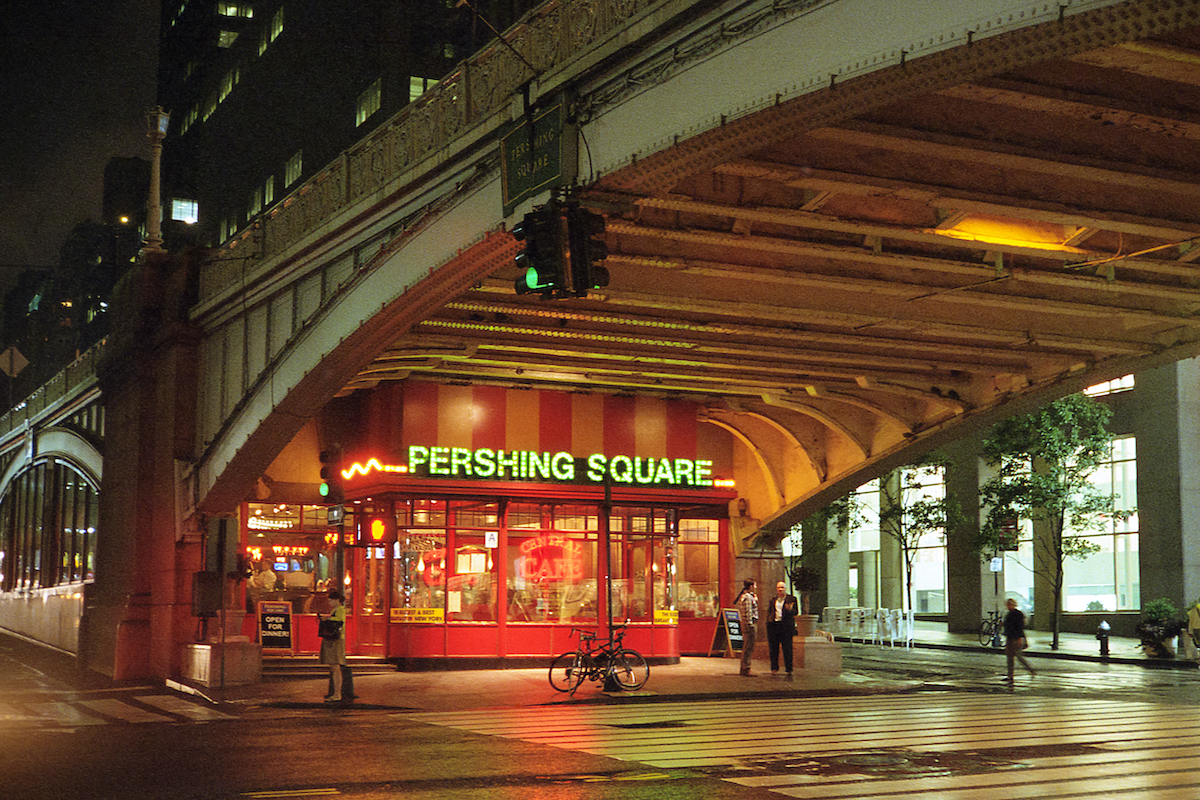 Un restaurante bien iluminado con letras verdes fluorescentes que dicen Pershing Square debajo de un paso elevado urbano