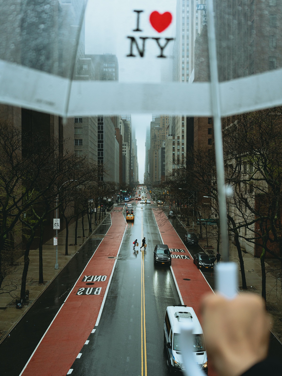 Una persona con un paraguas transparente que dice "I love NY" sobre un paisaje urbano lluvioso