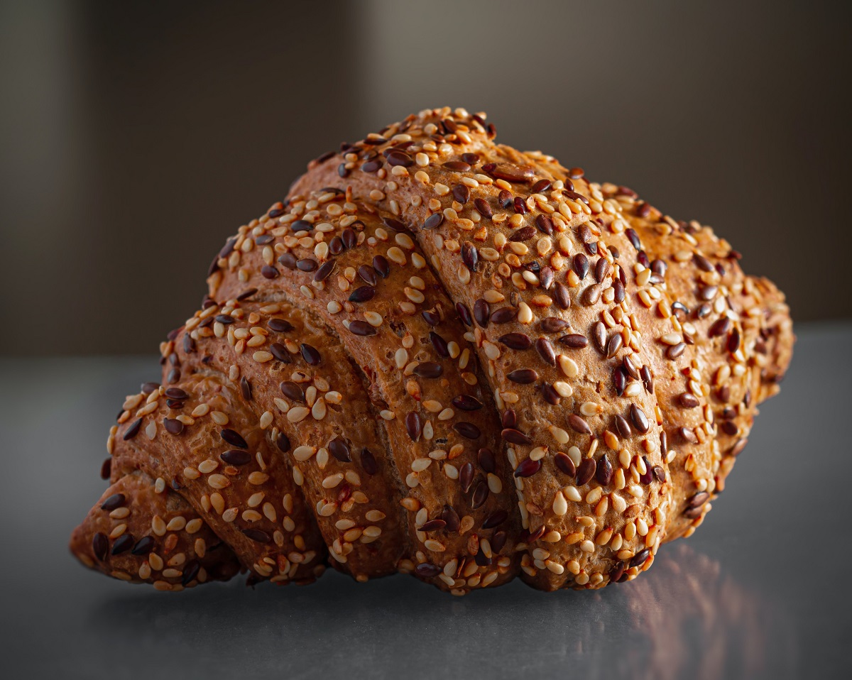 Un croissant regordete cubierto con semillas de diferentes colores sobre una superficie reflectante
