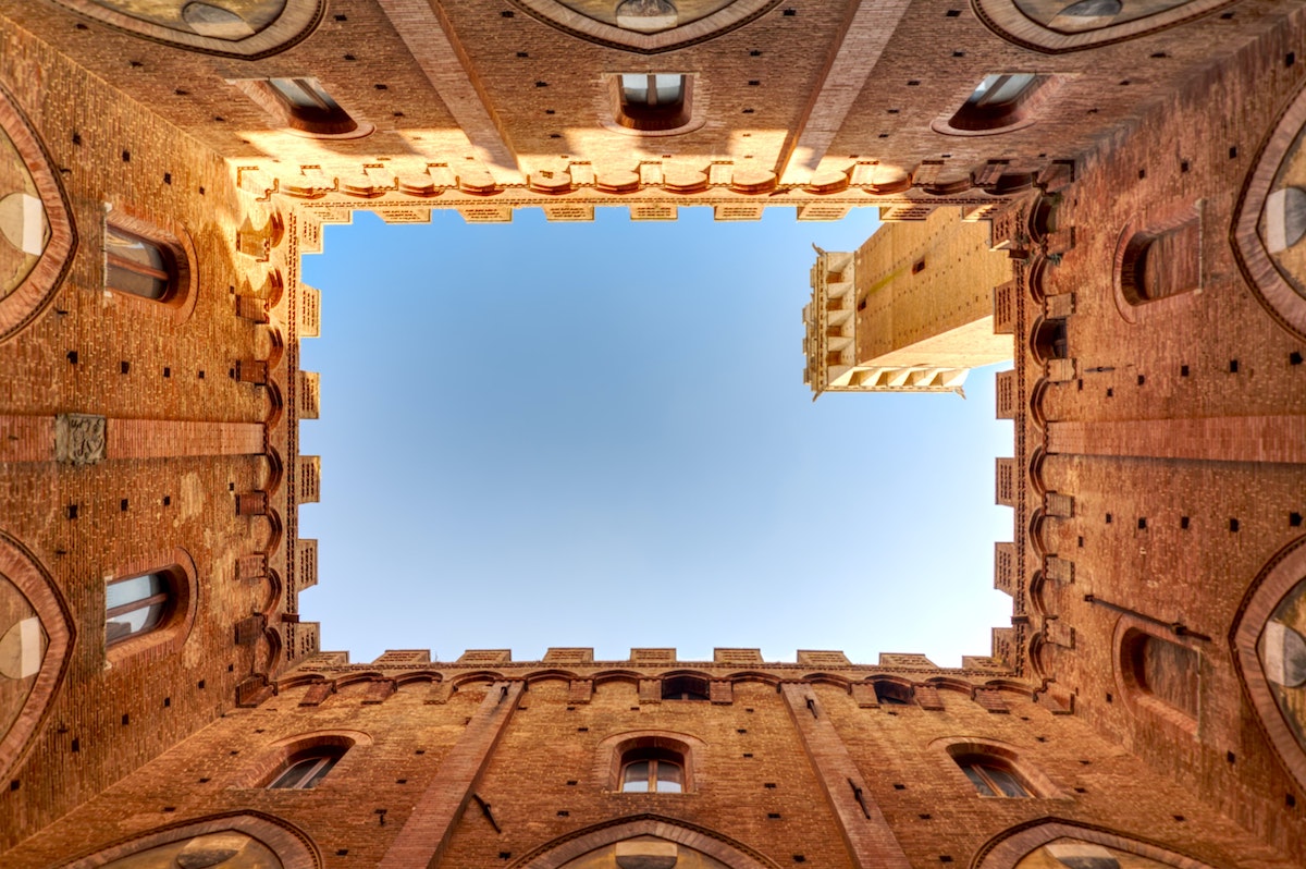 Vista del cielo tomada desde el interior de un patio rectangular con edificios medievales marrones y una torre visible en los bordes.