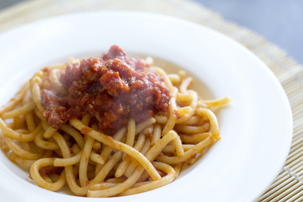Fideos gruesos tipo espagueti cubiertos con una salsa a base de salchicha, servidos en un plato blanco