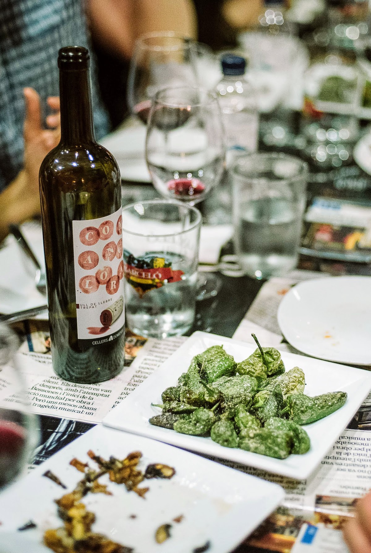 Pequeños pimientos verdes fritos en un plato blanco junto a una botella de vino