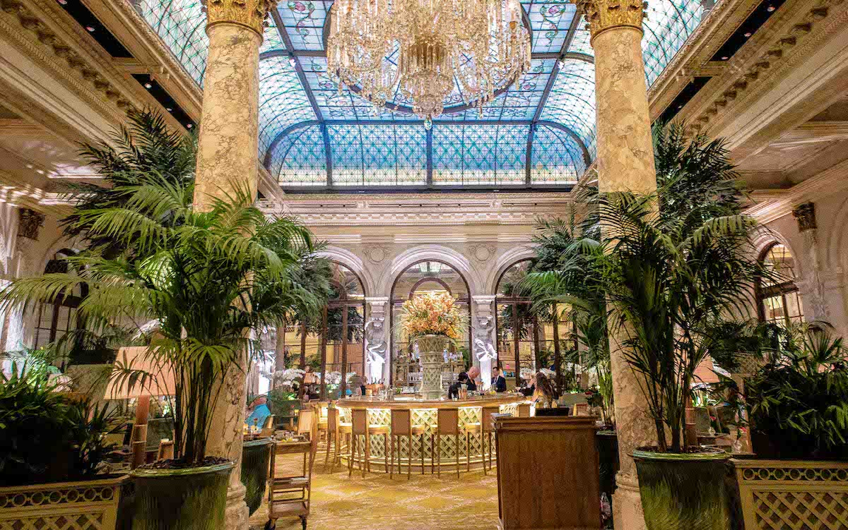 Bar de hotel de lujo adornado con una gran lámpara de araña y una entrada flanqueada por macetas con plantas y columnas de estilo griego