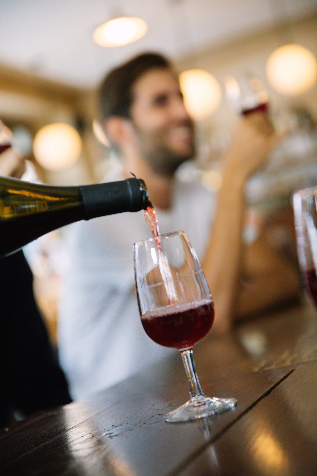 Cerca de una pequeña copa de vino tinto que se vierte con un hombre sonriente sosteniendo otra copa de vino en el fondo