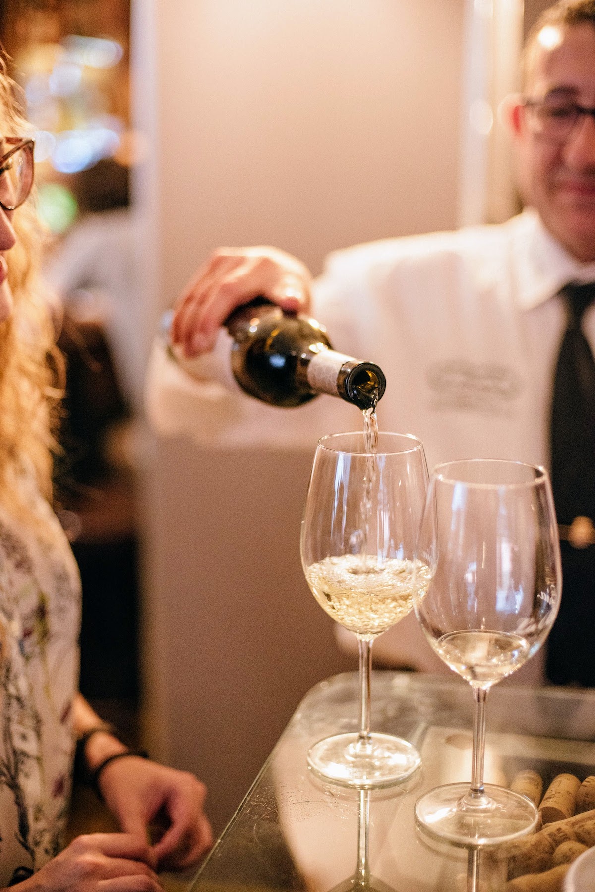 Un camarero con camisa blanca y corbata sirviendo dos copas de vino blanco