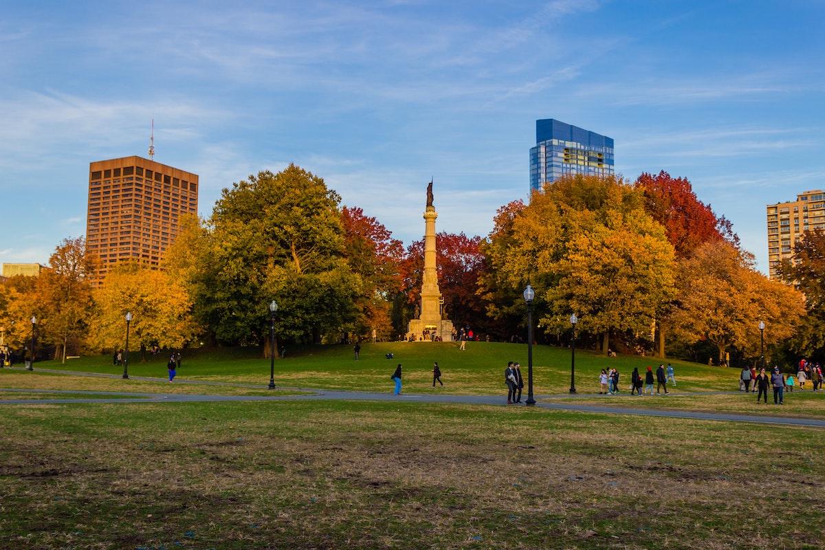 El paisaje de otoño de Boston Public Garden. La gente camina por senderos pavimentados, rodeados de árboles de colores vibrantes. Los rascacielos miran al fondo.