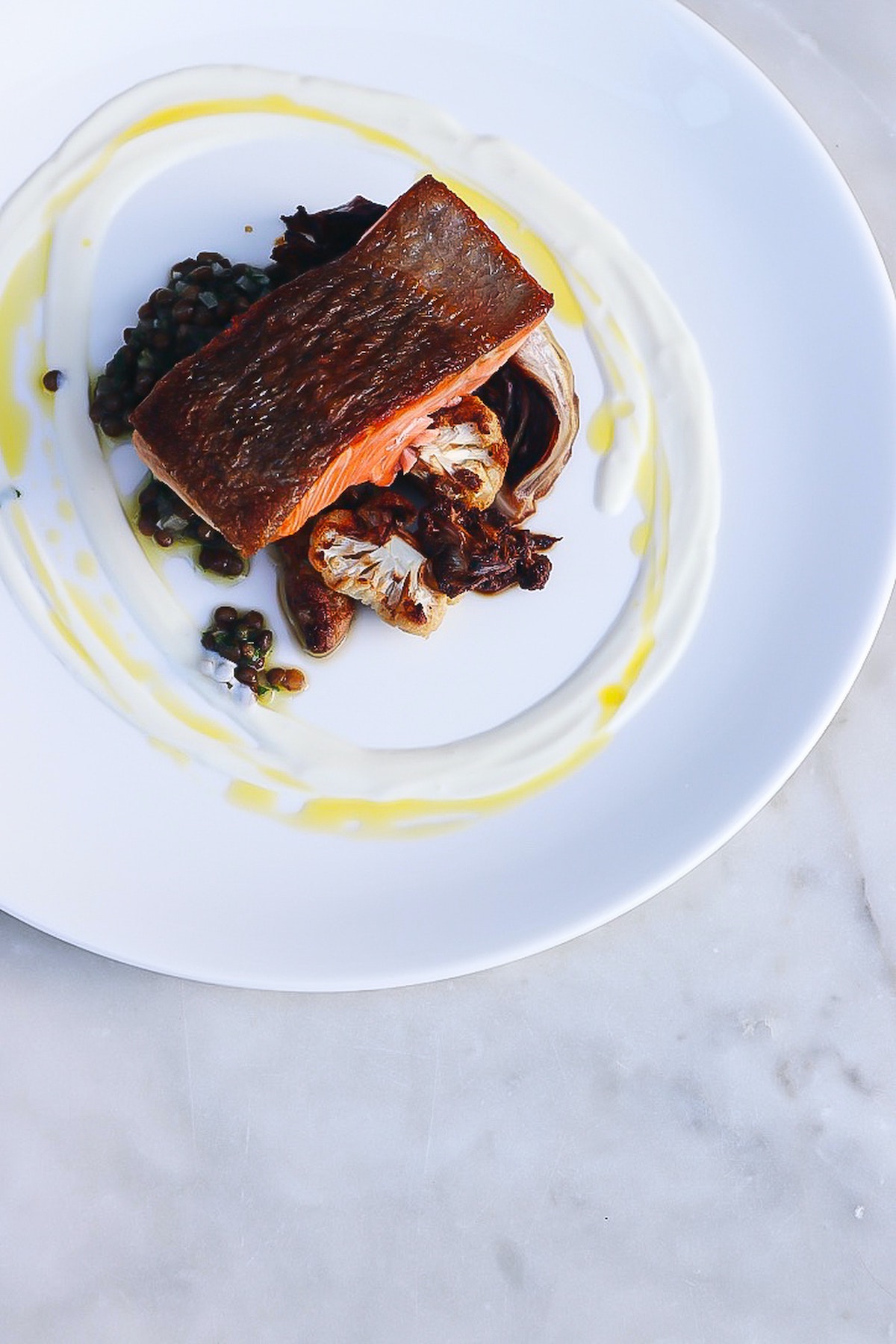 Fotografía cenital de filete de salmón ennegrecido y coliflor en un plato blanco ingeniosamente decorado con salsa blanca y amarilla