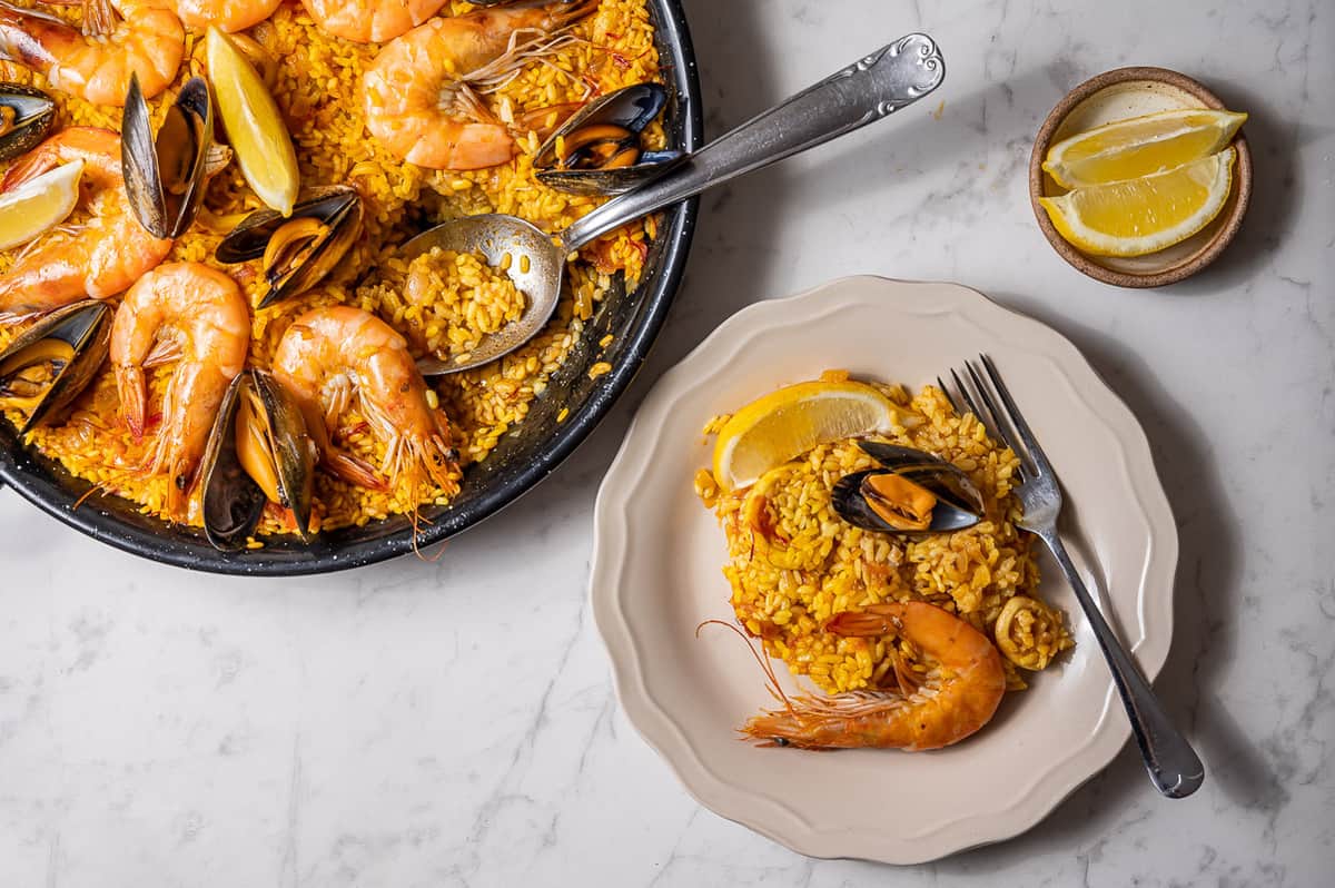 Fotografía cenital de una paella de marisco con una pequeña porción servida en un plato blanco.