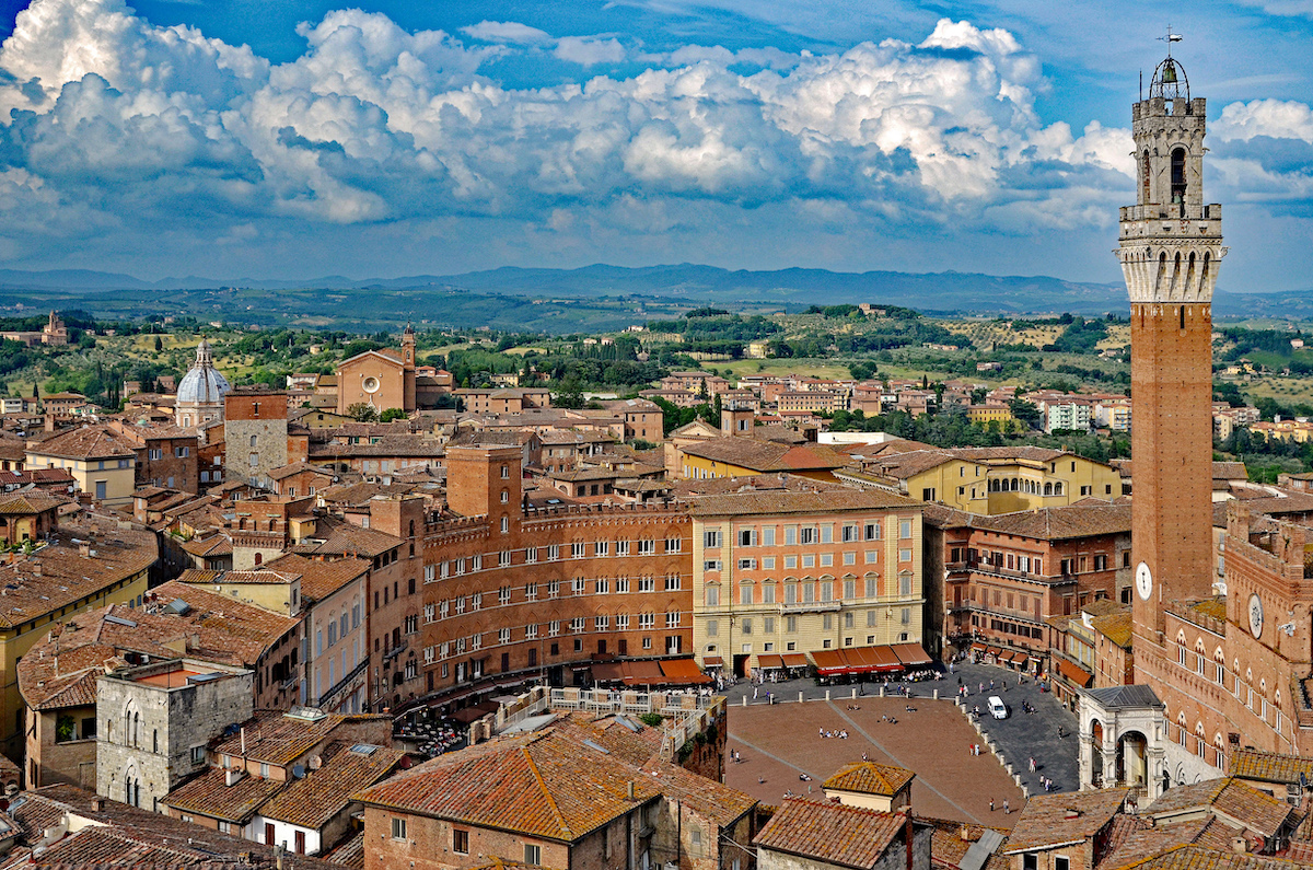 Vista aérea de Siena, Italia, con edificios marrones históricos y una torre prominente a la derecha, con el campo verde al fondo