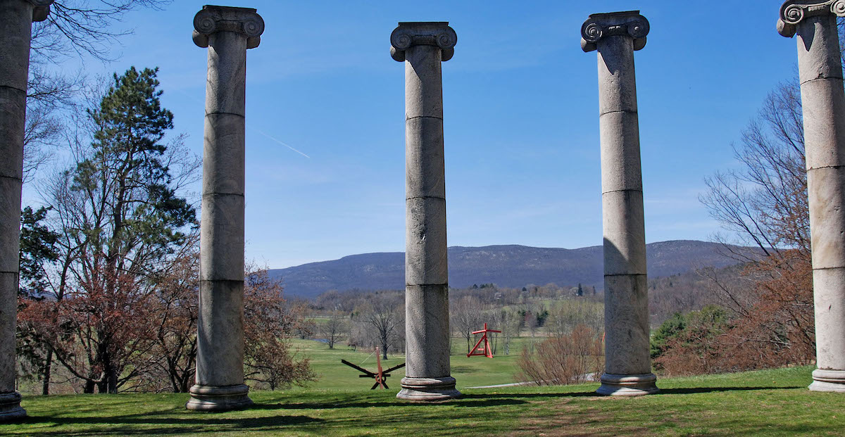 Cinco grandes columnas de piedra en un espacio abierto al aire libre y varias otras estatuas se pueden ver al fondo