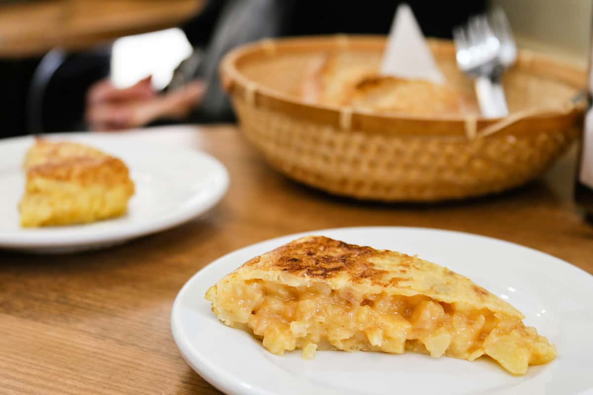 Dos rebanadas de tortilla de patata española en platos blancos separados con una cesta de pan integral en el fondo.