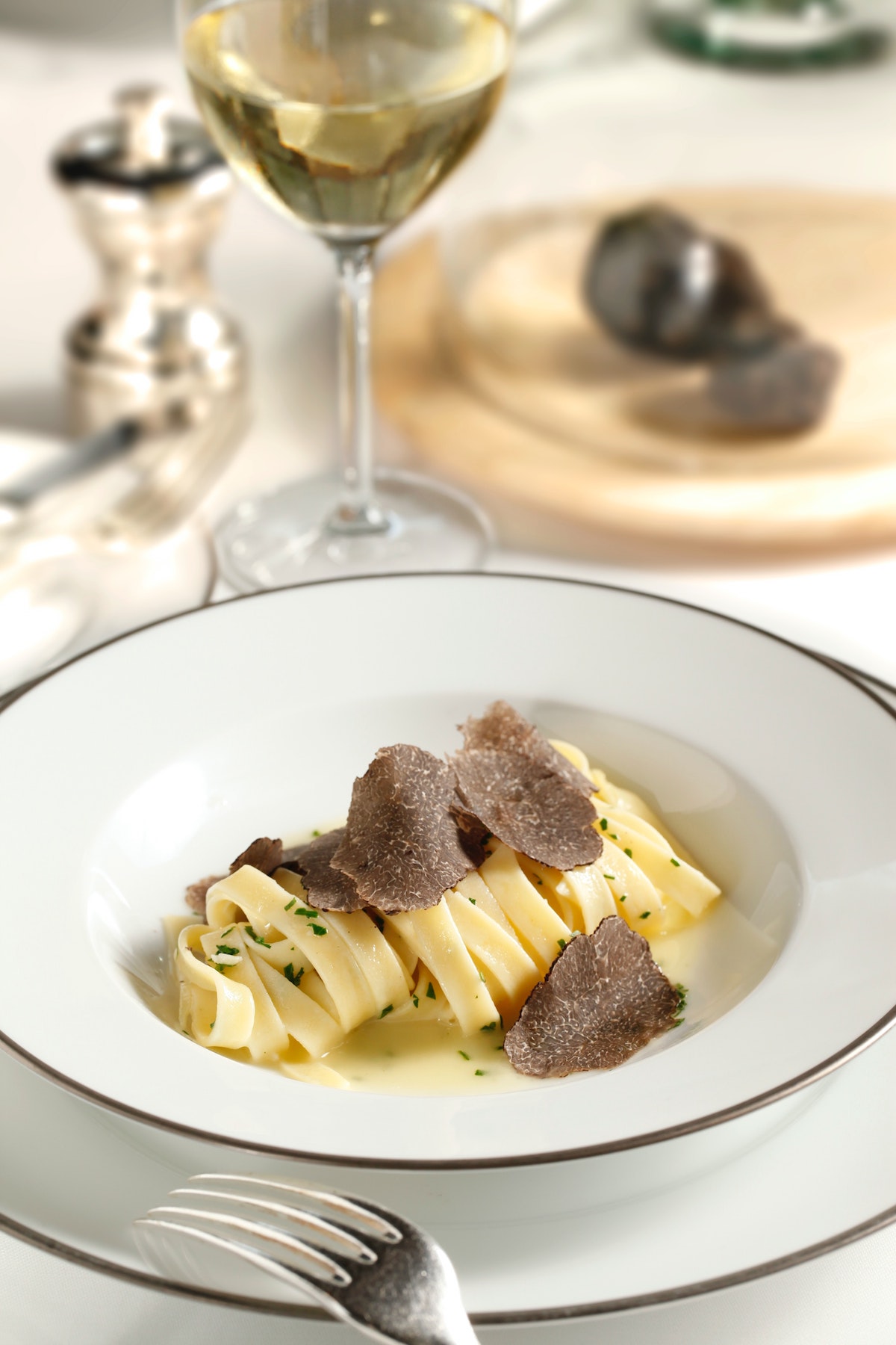 Un plato de pasta hecho con fideos planos y anchos y espolvoreado con trufas sobre una mesa junto a una copa de vino blanco.