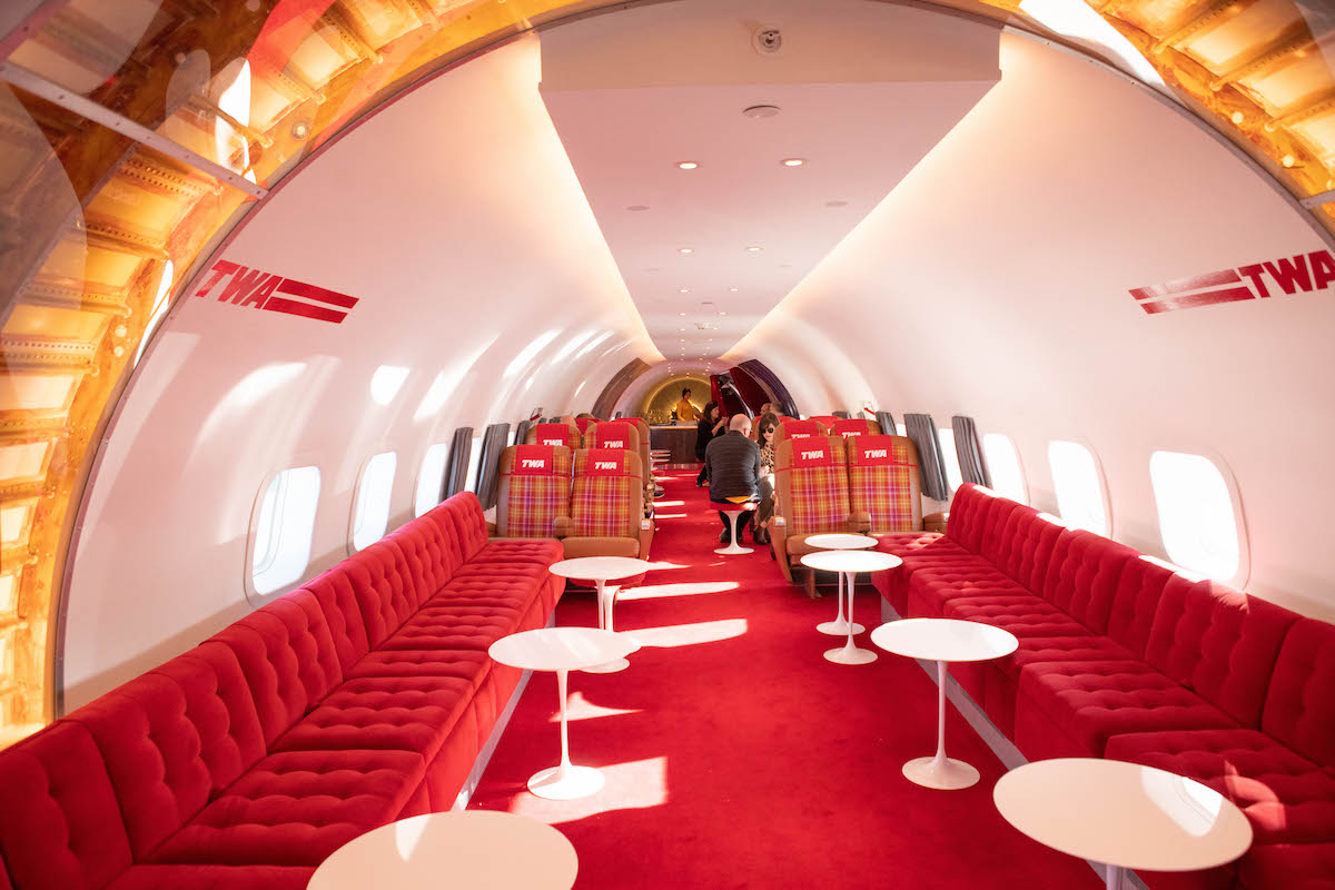 Restaurante dentro de un avión TWA restaurado, con alfombra roja y asientos de banco, pequeñas mesas blancas redondas y asientos de avión a cuadros en el fondo